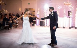 A bride and groom dance on a fog-covered floor in a decorated reception hall, surrounded by seated guests and elegant lighting.