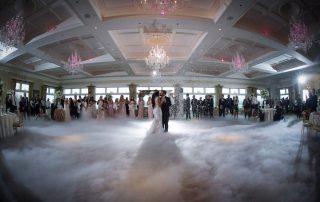 A bride and groom share their first dance in a grand ballroom filled with guests, chandeliers, and a low-lying fog effect on the dance floor.