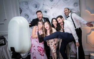 Six adults in formal attire pose making silly faces and gestures in front of a white floral backdrop at a photo booth.
