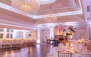 Elegant ballroom set for an event with round tables, gold chairs, floral centerpieces, chandeliers, and candles, featuring ornate ceilings and a lit fireplace.