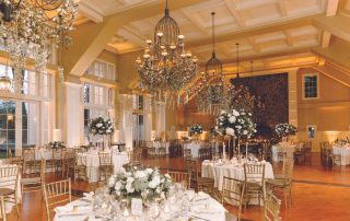 Elegant banquet hall with round tables set for an event, featuring white floral centerpieces, gold chairs, and large chandeliers under a high ceiling.