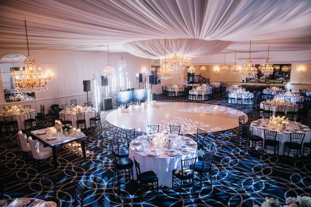 The Gramercy at Lakeside Manor in Hazlet NJ has an elegant ballroom set for an event with round tables, black chairs, a large empty dance floor, chandeliers, draped ceiling, and soft lighting.
