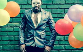 A person wearing a gray suit and a white mask stands against a green brick wall surrounded by colorful balloons.