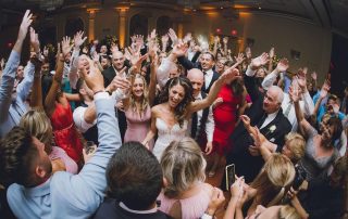 A bride and groom celebrate on a crowded dance floor, surrounded by guests with raised hands in a festive ballroom setting.