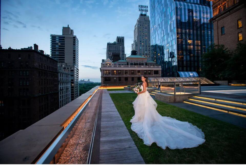 A bride in a white wedding gown stands on a rooftop garden at dusk, holding a bouquet, with city skyscrapers and buildings in the background.