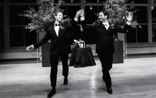 Two people in tuxedos walk hand in hand on a wooden floor, raising their arms in celebration, with large floral arrangements and a table in the background.