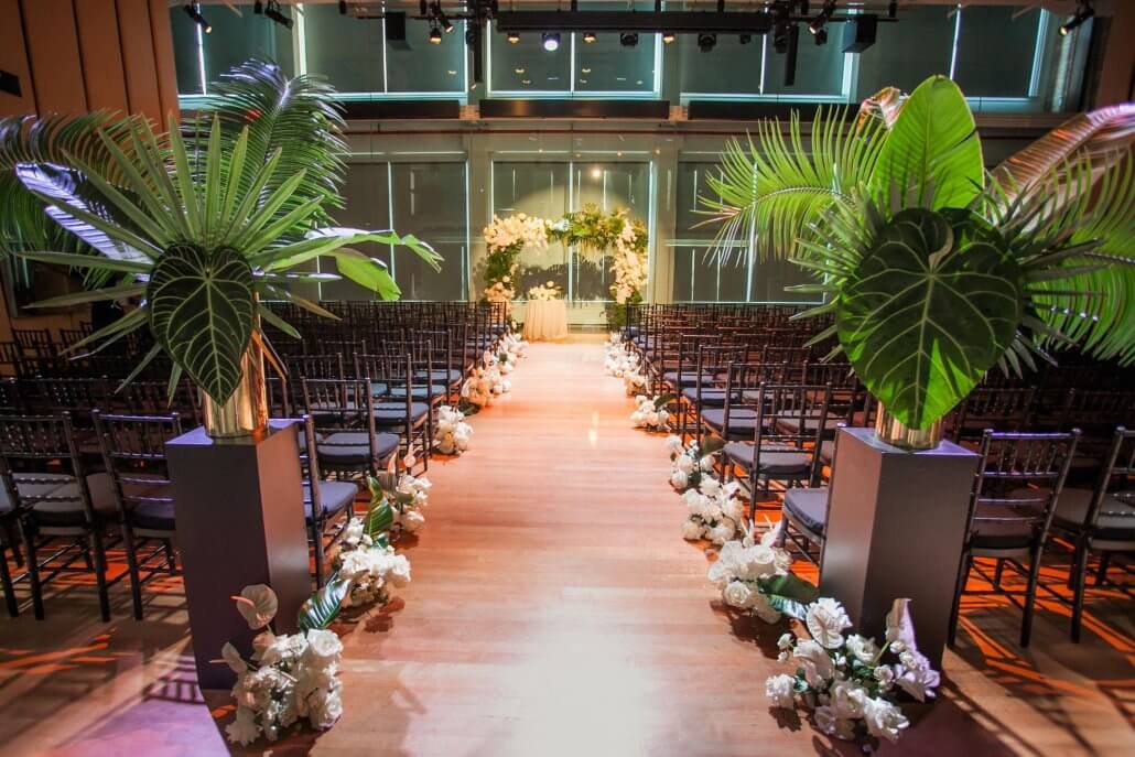 A wedding ceremony venue with rows of chairs, a decorated aisle lined with flowers, and a floral arch at the front. Large green plants are placed at the entrance to the aisle.