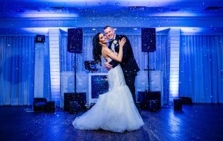 A bride and groom in formal attire embrace and smile while dancing on a decorated indoor stage with artificial snow falling around them.