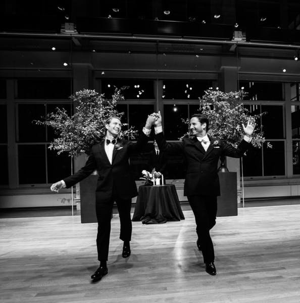 Two men in tuxedos walk hand-in-hand, raising their arms in celebration on a dance floor, with a table and decorative trees in the background.