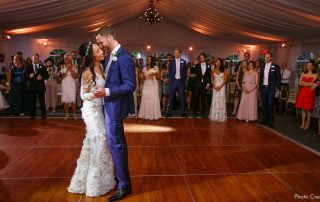 A bride and groom share their first dance on a wooden floor under a tent, surrounded by wedding guests watching and smiling.