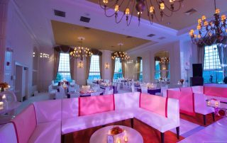 Large elegant banquet hall with pink and white sofas, round tables, chandeliers, and tall windows set up for an event, with ambient lighting.