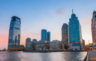 Skyline view of modern high-rise buildings along a waterfront at sunset, with reflections on the water and lights beginning to illuminate the city.