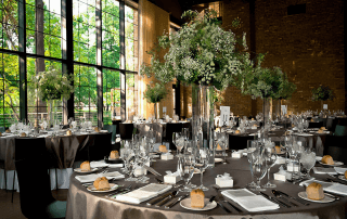 Elegant banquet hall with round tables set for an event, featuring tall floral centerpieces, glassware, plates, and bread rolls by large windows with greenery outside.