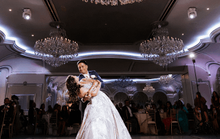 A bride and groom dance under large chandeliers in a ballroom, with wedding guests seated at tables in the background.