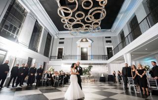 A bride and groom share their first dance in a grand ballroom with high ceilings, checkered floors, and guests watching from the sides.