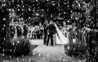 A wedding ceremony takes place outdoors, with the couple exchanging vows in front of guests; the scene is in black and white with decorative lights in the foreground.