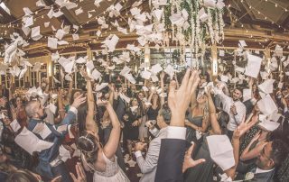A large group of people in formal attire celebrate indoors, throwing papers in the air under hanging lights and greenery.