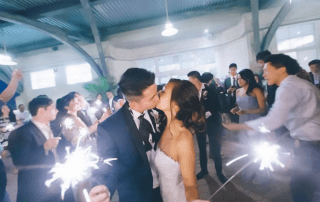 A bride and groom kiss while holding sparklers, surrounded by guests celebrating indoors.