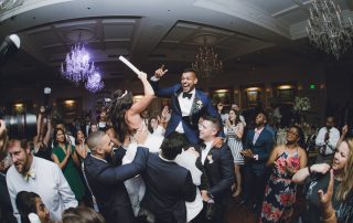 A bride and groom are lifted in the air by guests on a crowded dance floor at a wedding reception, with people cheering and celebrating around them.