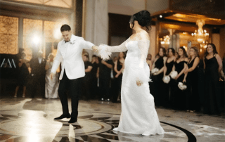 A bride and groom dance on a marble floor in a grand hall, while a group of bridesmaids in black dresses watch from the background.