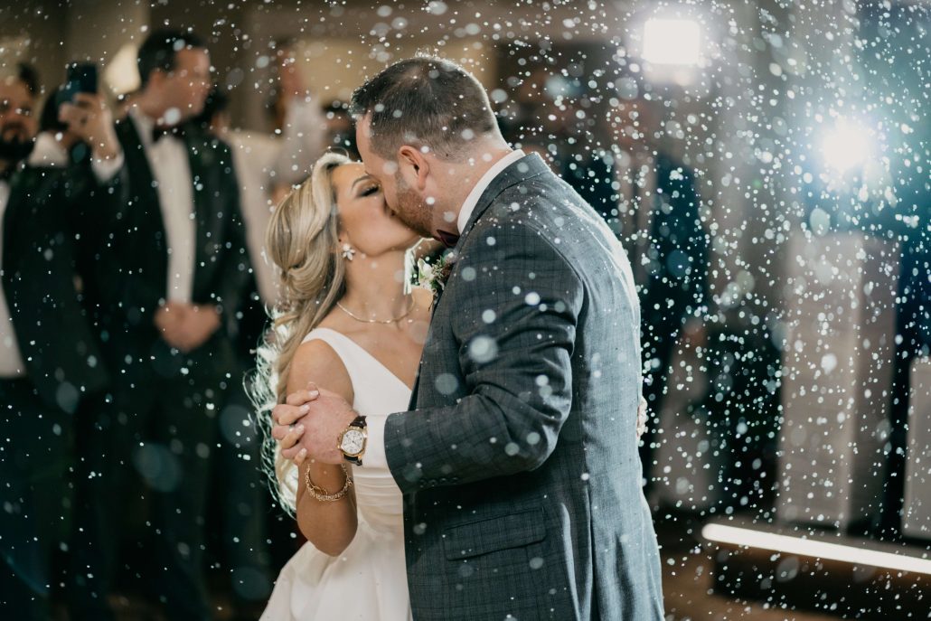 A bride and groom share a kiss on the dance floor as artificial snow falls around them, with guests and camera flashes visible in the background.