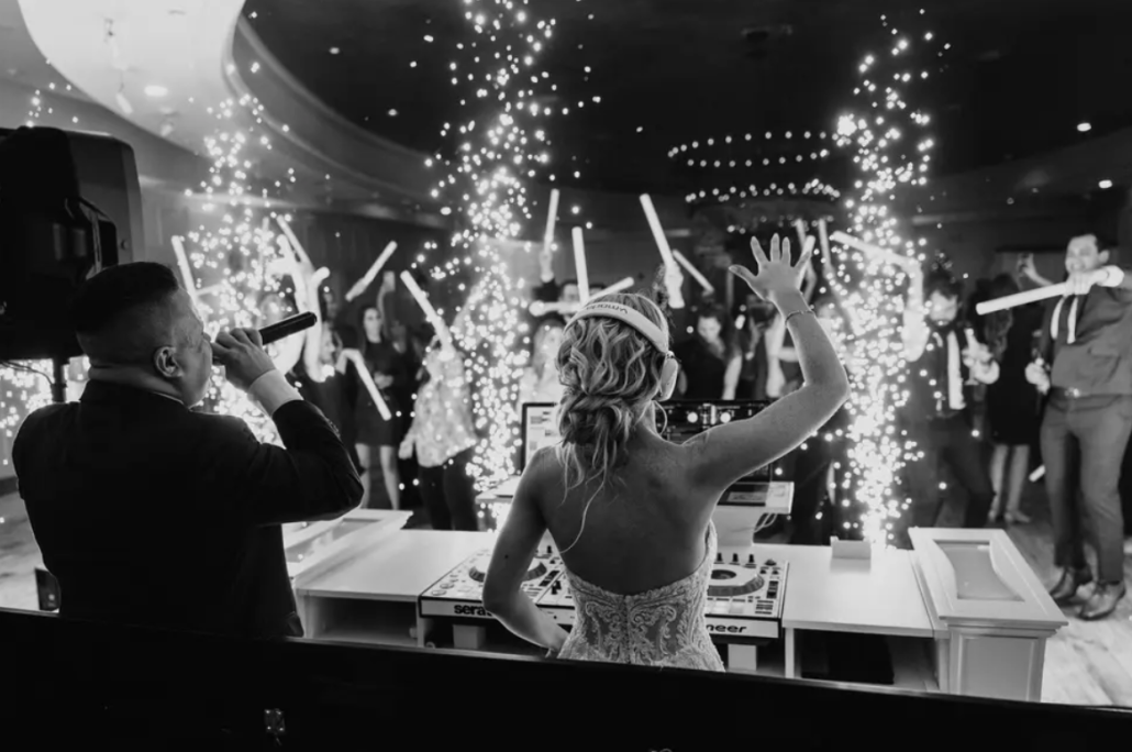 A bride faces a dancing crowd as a man sings into a microphone; indoor fireworks and light sticks add to the celebration atmosphere.