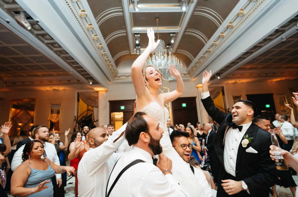 A bride is lifted on a chair during a wedding reception while guests around her cheer and dance in a grand ballroom.