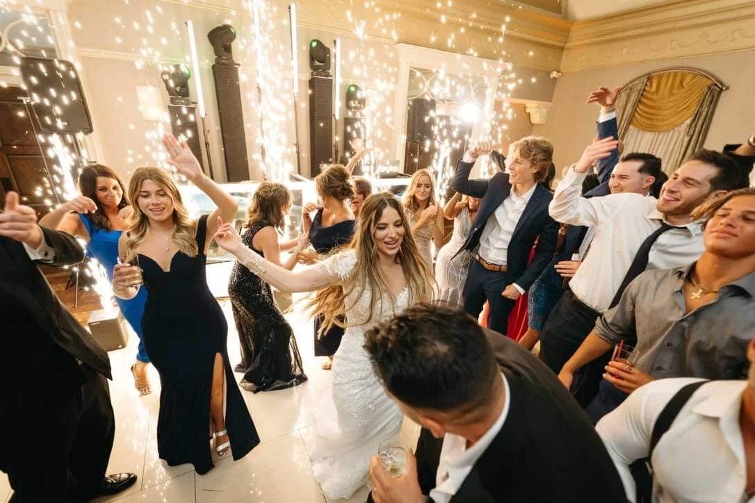 bride-and-groom-dancing-in-front-of-cold-sparks A bride and guests dance and celebrate at a wedding reception with bright sparklers and lively atmosphere in a decorated indoor venue.