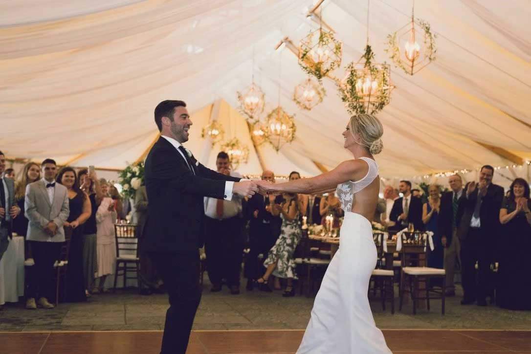 bride-and-groom-dancing-in-reception-tent-sce A bride and groom share their first dance in a decorated tent, surrounded by guests watching and smiling.