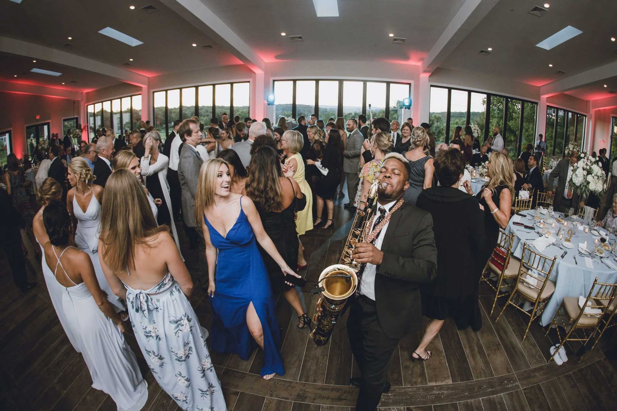 saxophone-player-at-nj-wedding-sce-event-group A crowded indoor event with people dancing and a musician playing the saxophone among tables set for a formal gathering.