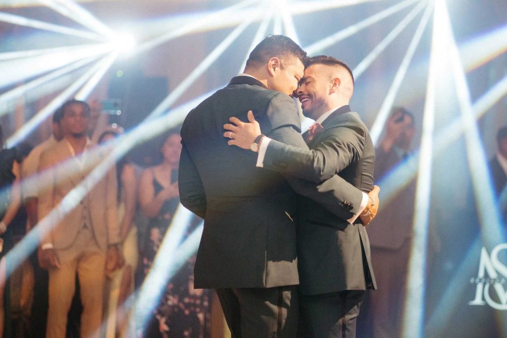 Two men in suits share a close dance on a lit-up dance floor, surrounded by guests and beams of light.