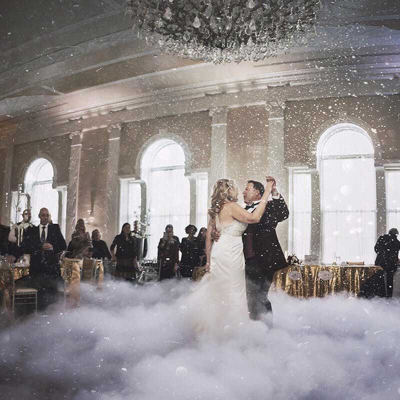 A bride and groom share their first dance in a ballroom filled with low-lying fog, surrounded by guests and elegant decor.