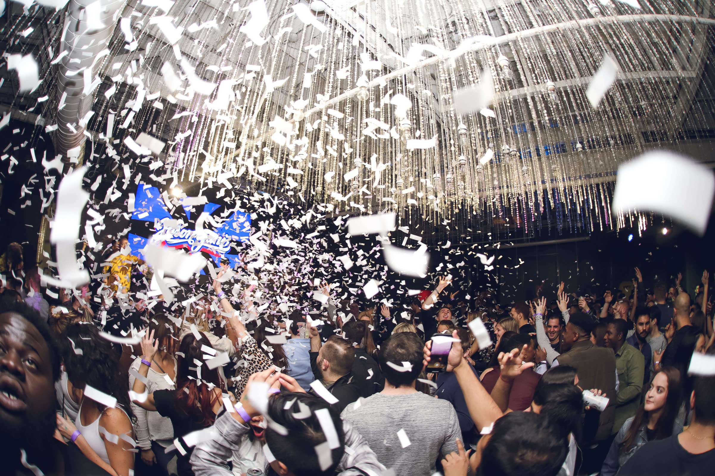 A crowded indoor party with people celebrating and confetti falling from the ceiling under bright lights.