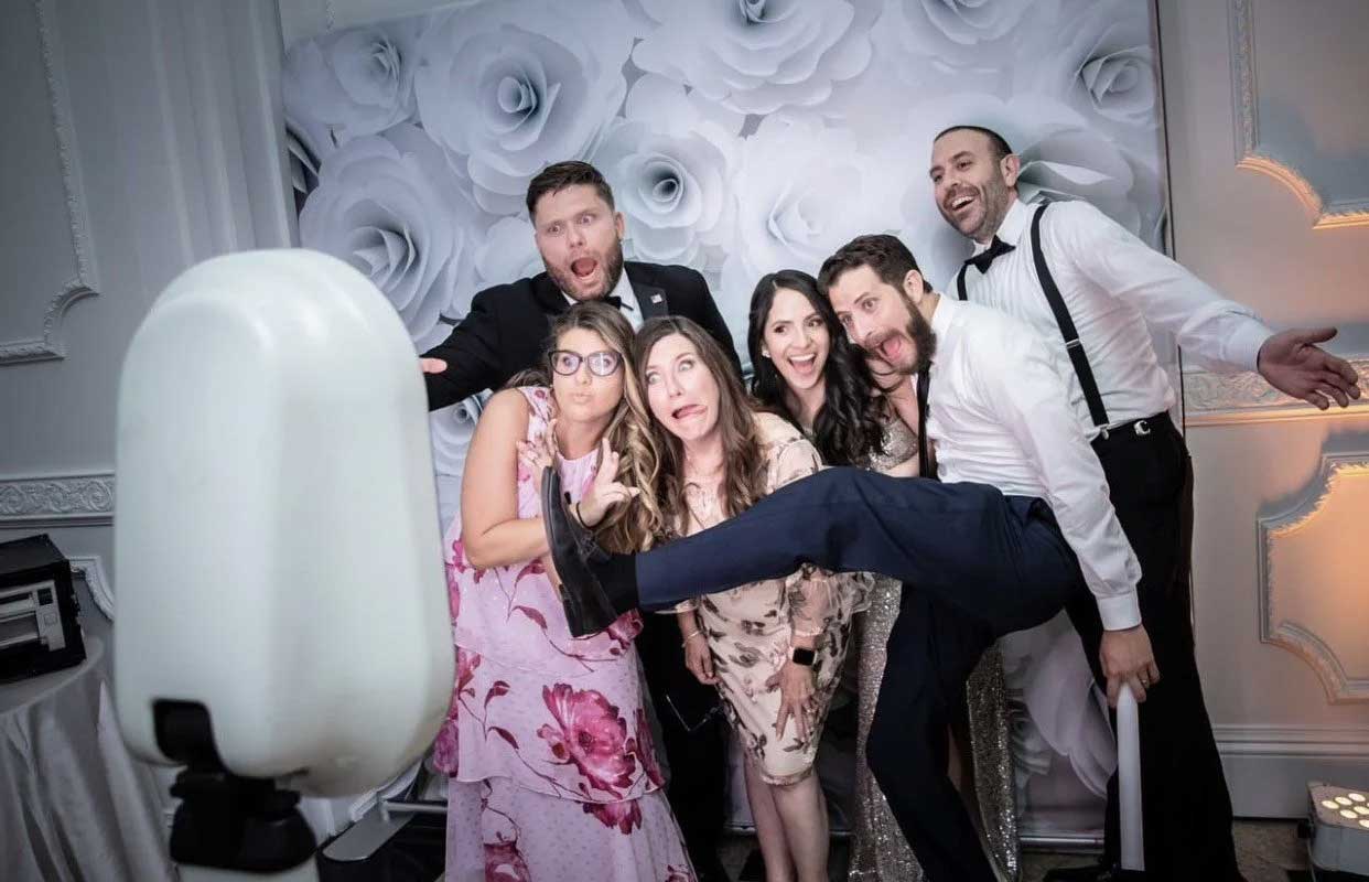 Six adults pose playfully with exaggerated expressions in front of a floral backdrop at a photo booth, with one man lifting his leg across the group.