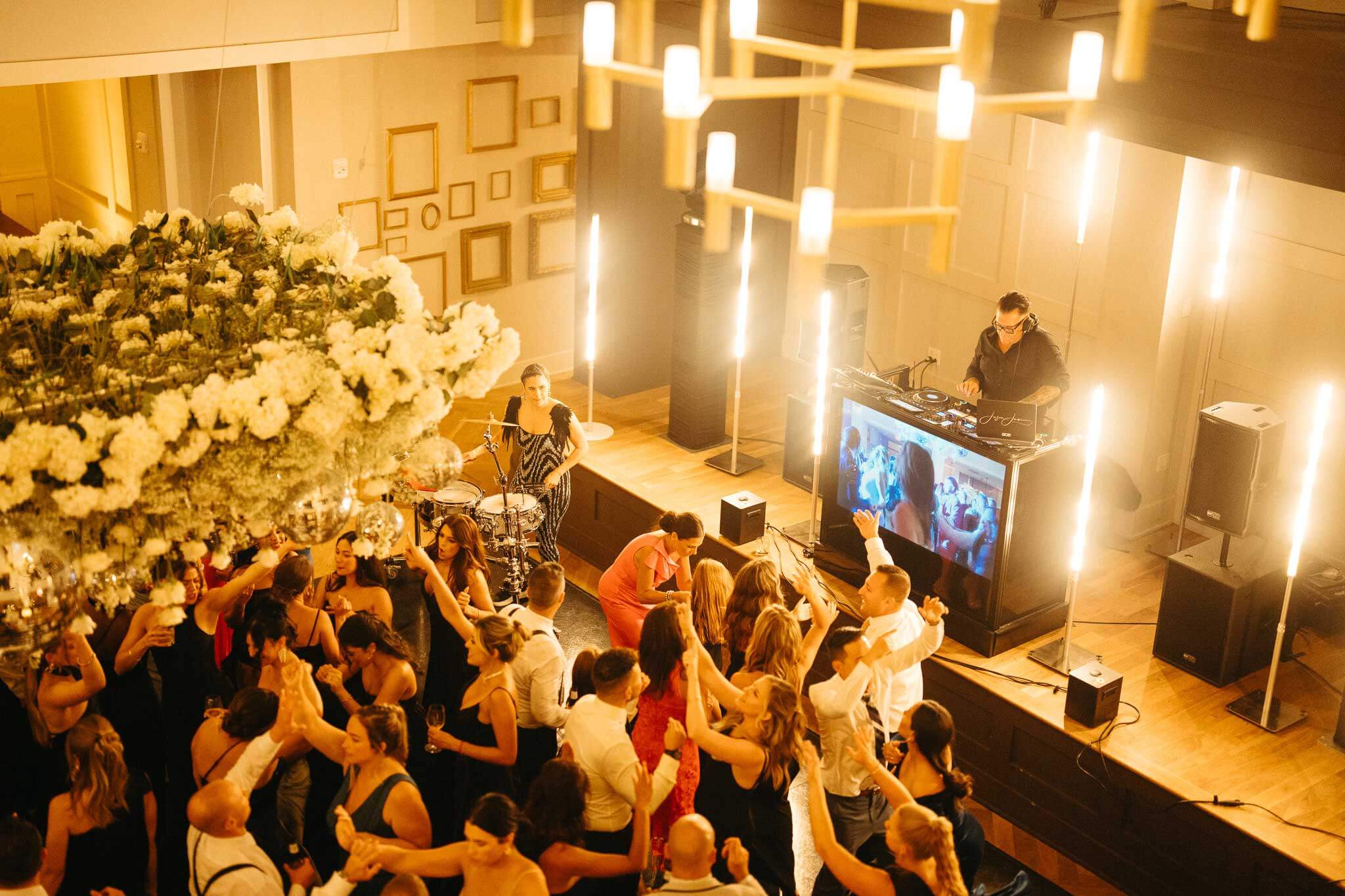 A crowd of people dance in a warmly lit indoor venue with a DJ performing on stage and a drummer beside large floral decorations.