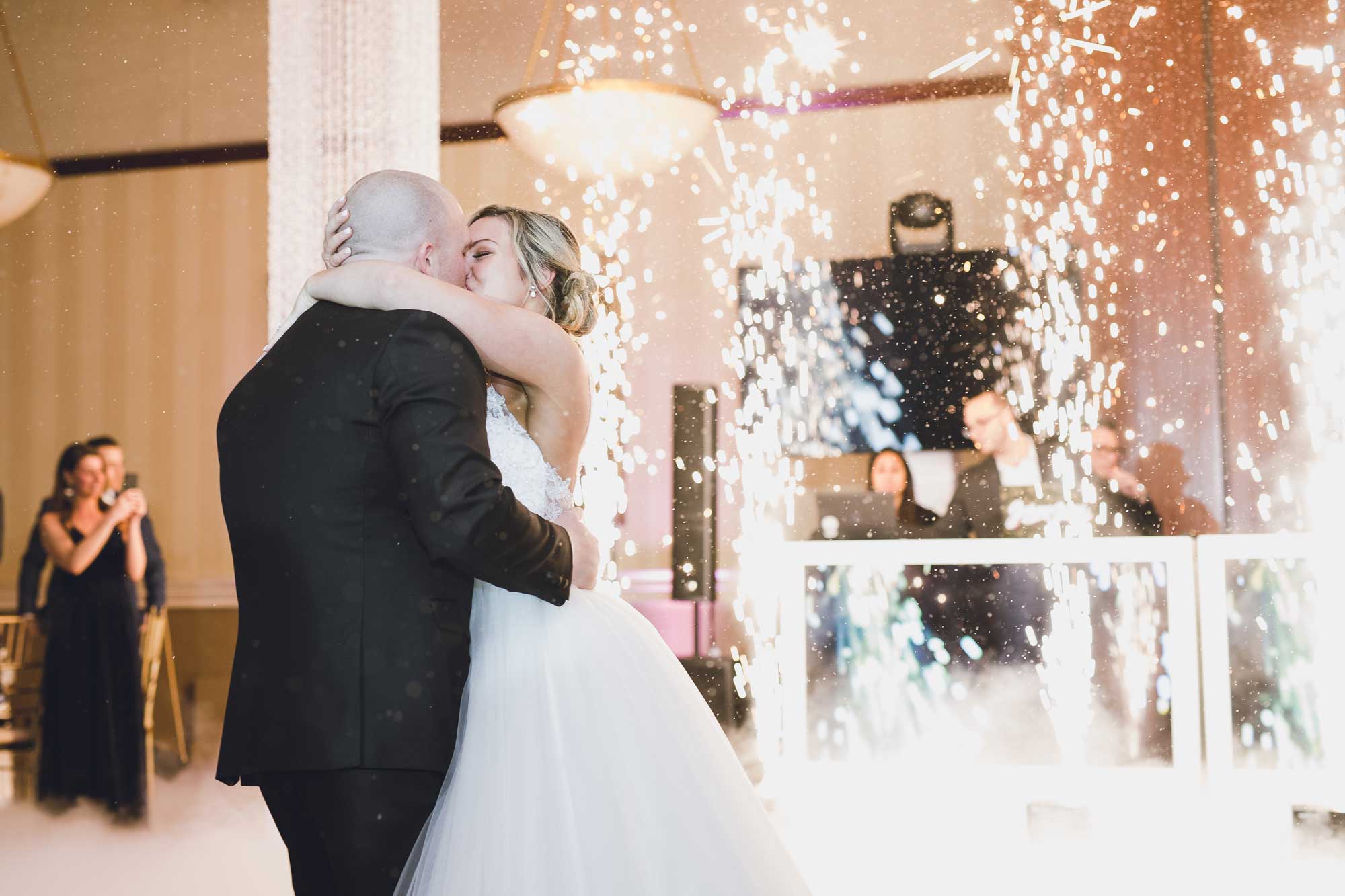 A bride and groom share a kiss on the dance floor surrounded by fireworks, with guests and DJ booth visible in the background.