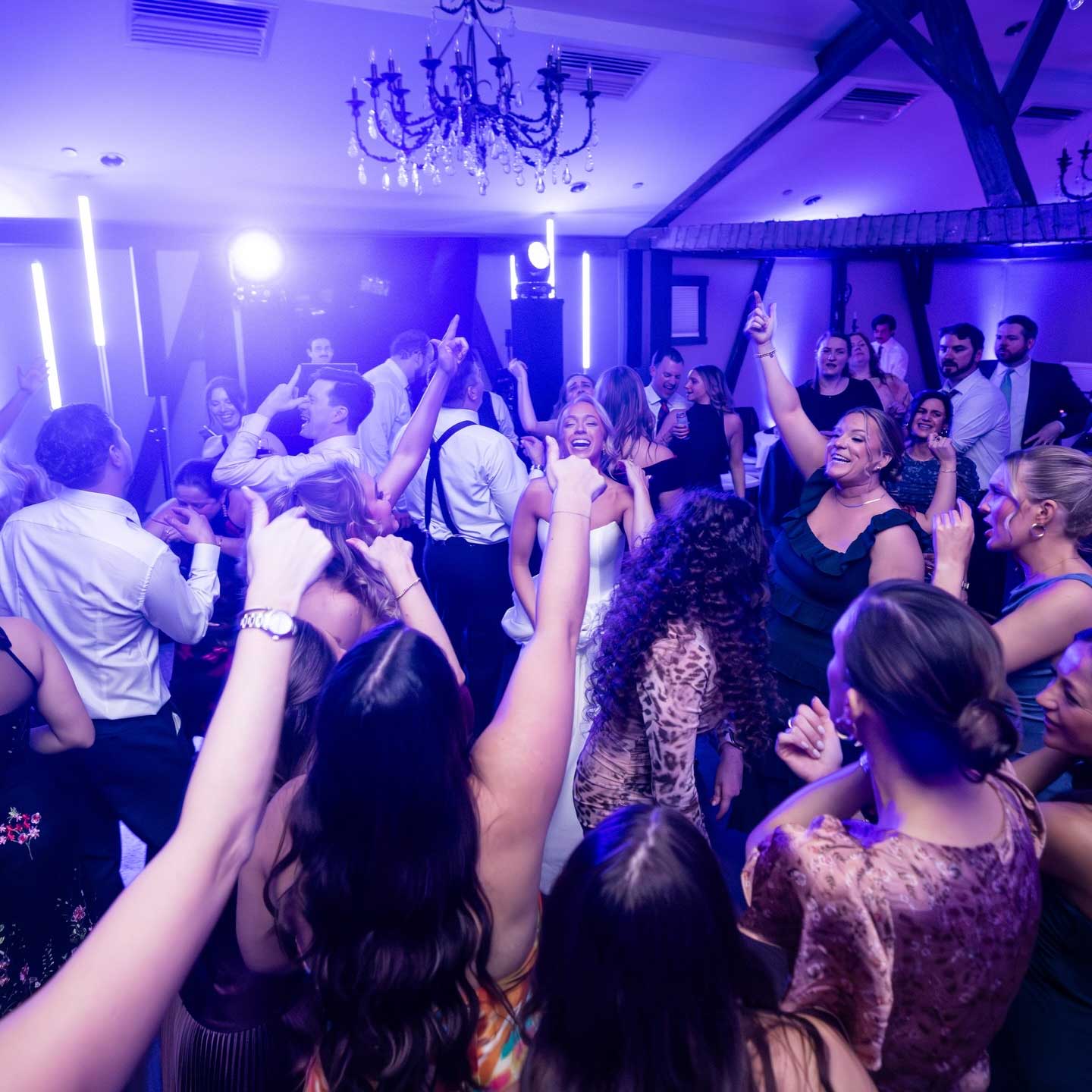 A group of people are dancing and celebrating in a dimly lit room with purple lighting and a chandelier overhead.