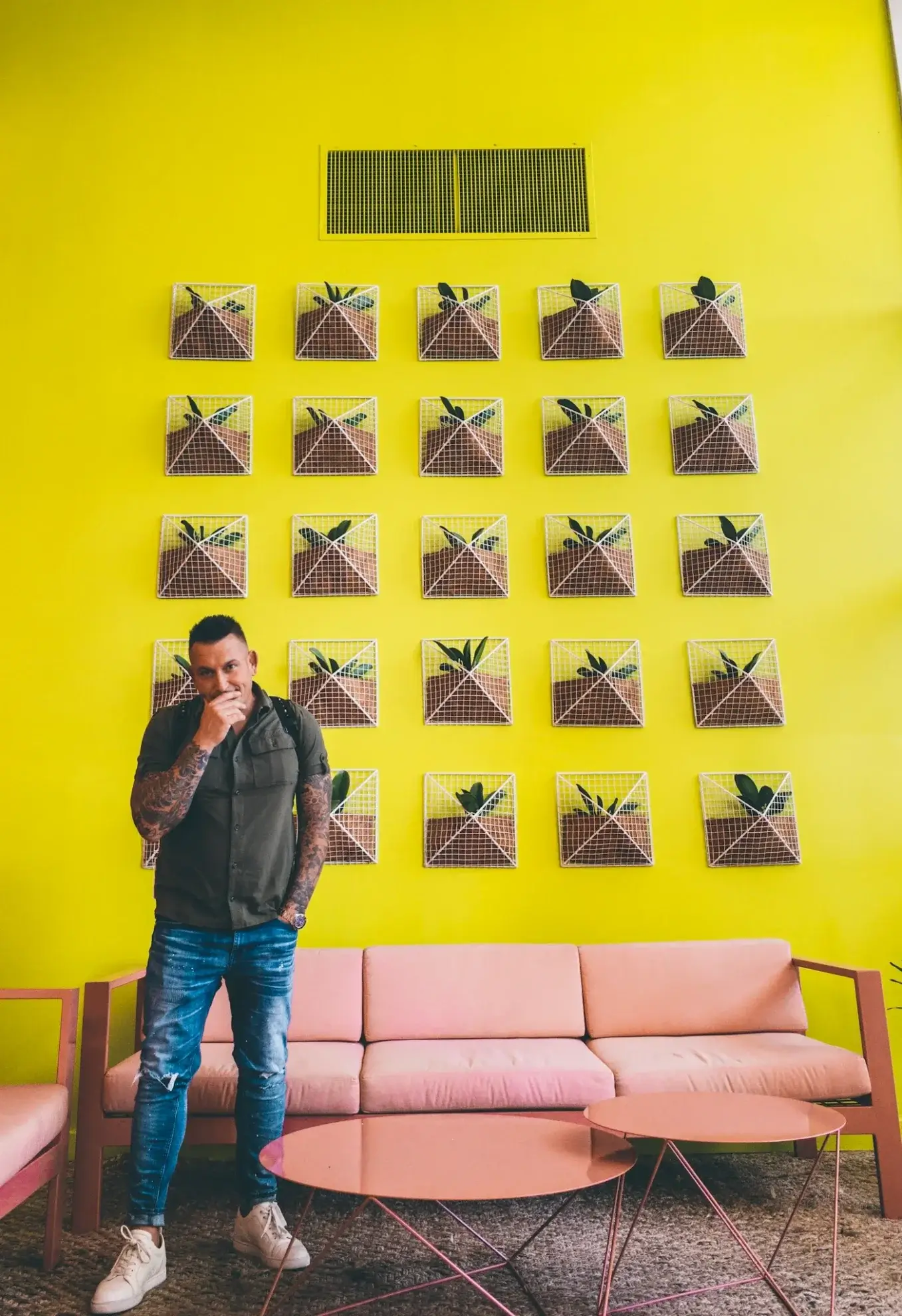 A man stands in front of a yellow wall with a grid of small wall planters above a pink sofa and matching tables.