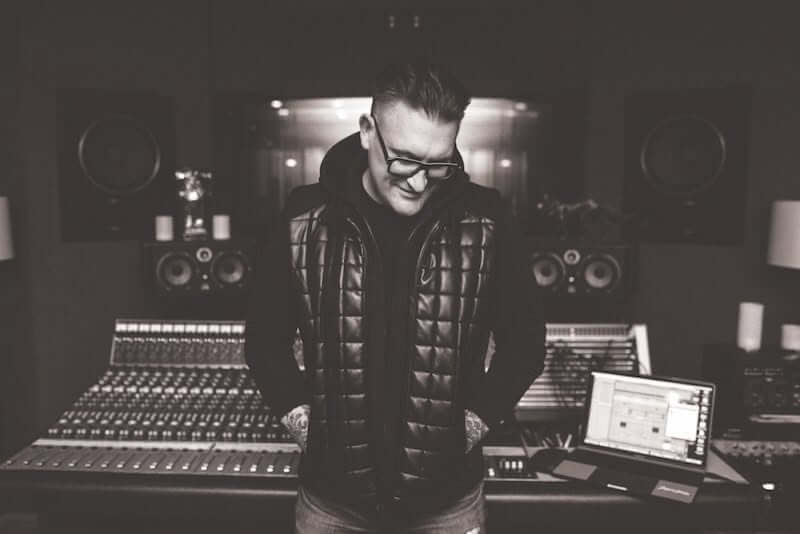 A person wearing glasses and a padded vest stands in front of a large audio mixing console and studio equipment in a recording studio.