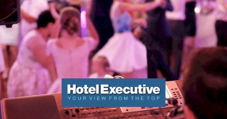A blurred group of people in formal attire dance at an event, viewed from behind a sound mixing console, with the "Hotel Executive" logo overlaid.
