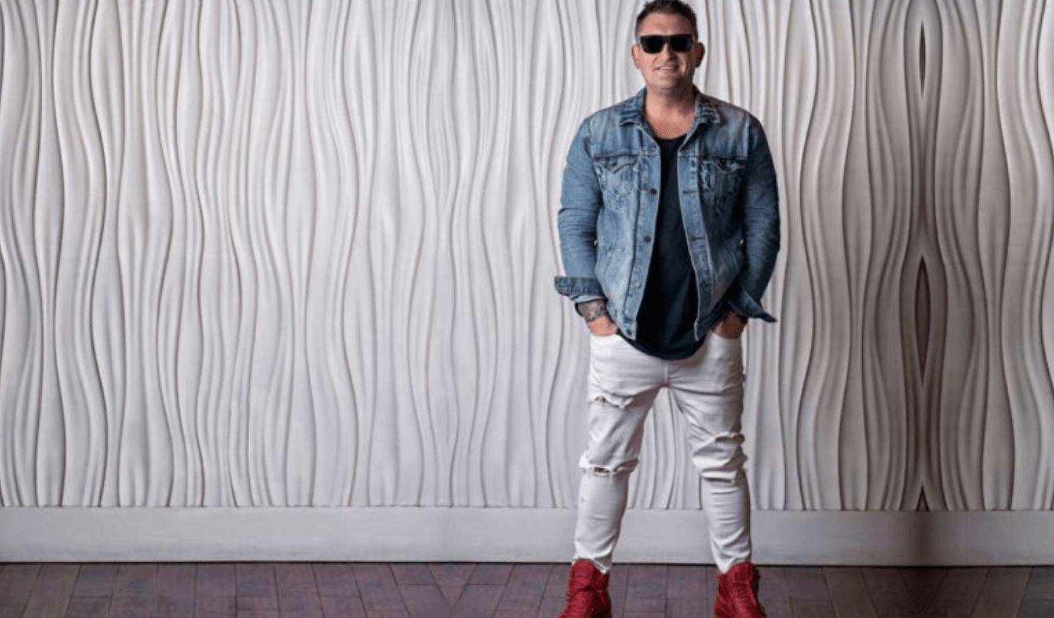 Man wearing sunglasses, a denim jacket, black shirt, white ripped jeans, and red sneakers stands in front of a wavy textured white wall on a wooden floor.