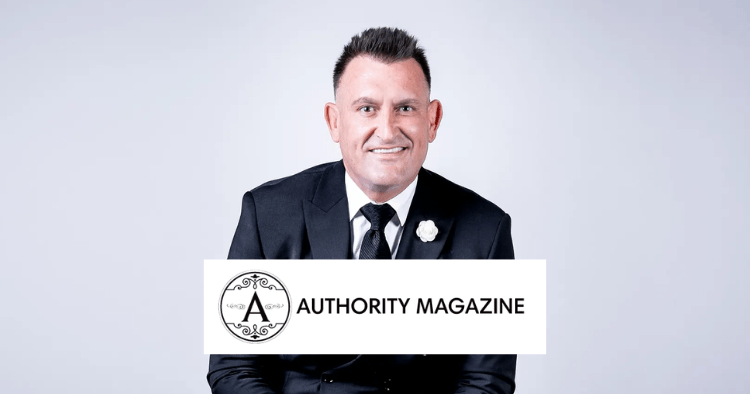 Man in a black suit with a white flower pin sitting and smiling, with the Authority Magazine logo in front of him.