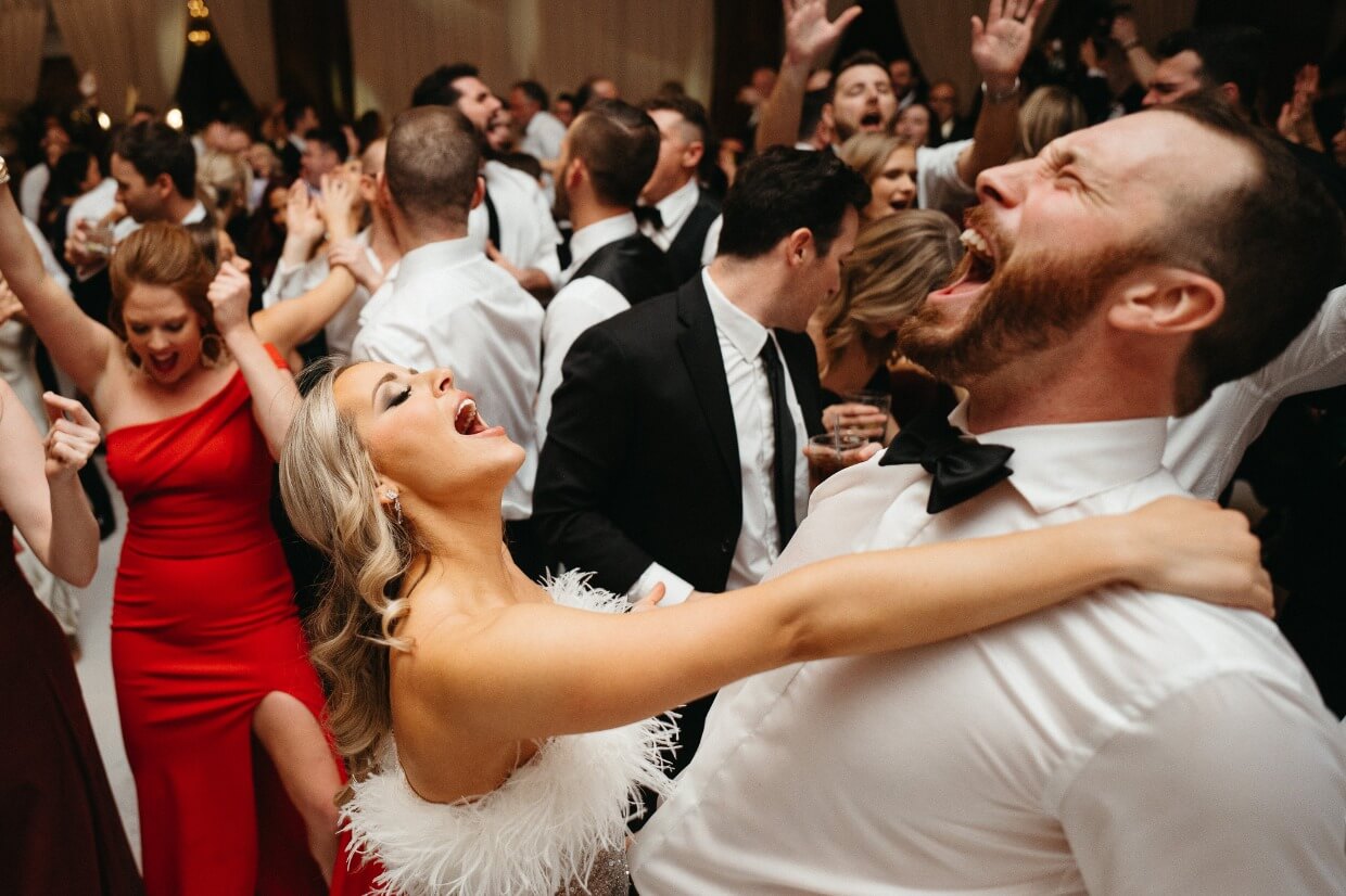 A group of people dancing and celebrating at a formal event, with two individuals in the foreground laughing and holding each other.