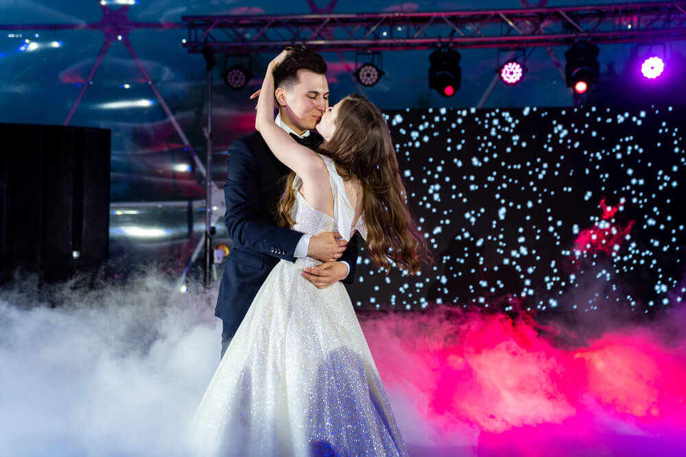 A couple in formal attire shares a romantic dance on a fog-covered floor with stage lights and a starry backdrop behind them.