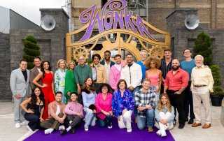 A group of people poses and smiles together on a purple carpet in front of a large decorative sign that reads “Wonka.”.