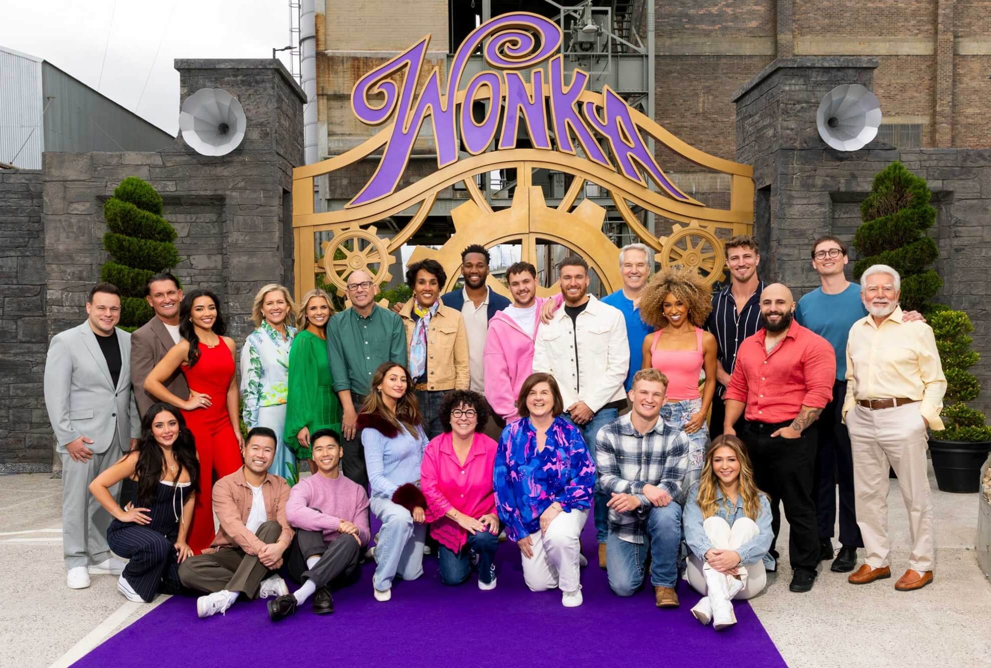 A group of people poses and smiles together on a purple carpet in front of a large decorative sign that reads “Wonka.”.
