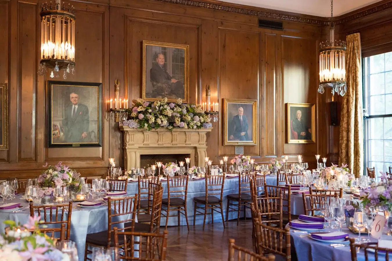 Formal dining room with round tables set for an event, floral centerpieces, wood-paneled walls, chandeliers, and portraits hanging above a fireplace.