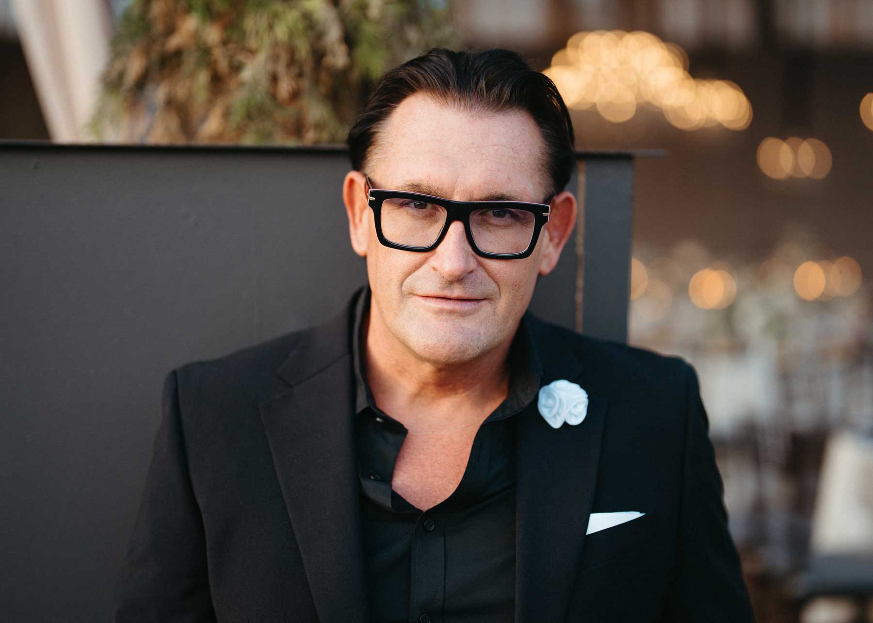 Man with dark hair and glasses wearing a black suit jacket, black shirt, and white flower pin, standing in front of a blurred background with lights.