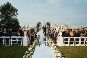 A bride and groom stand at an outdoor altar, exchanging vows. Guests are seated on either side of a flower-lined aisle, with a city skyline visible in the background.