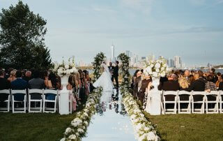 A bride and groom stand at an outdoor altar, exchanging vows. Guests are seated on either side of a flower-lined aisle, with a city skyline visible in the background.